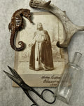 Vintage sepia-toned portrait print of a bearded man in historical clothing, styled with a seahorse, antler, scissors, and a small glass bottle on a textured lace surface.